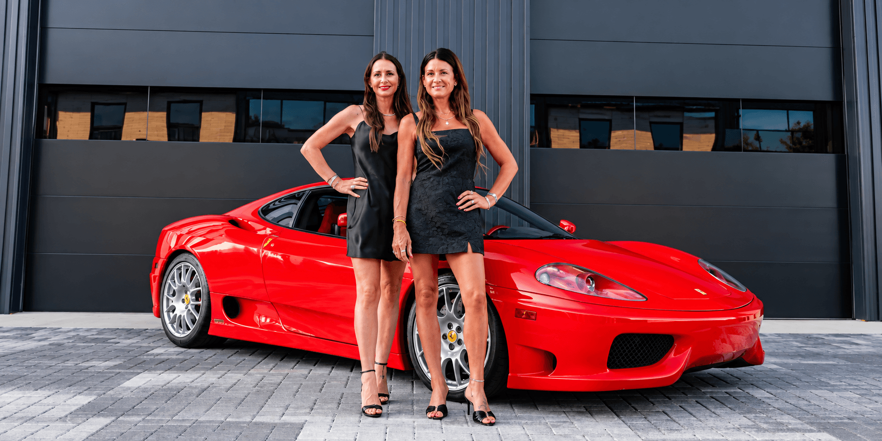 Two women in black dresses pose in front of a red sports car.