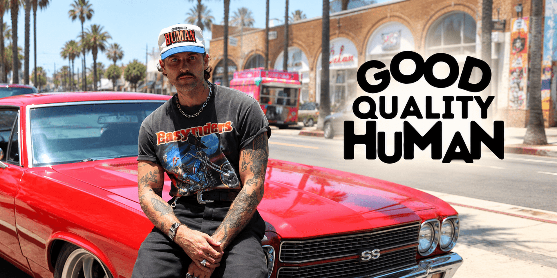 A tattooed man in a vintage t-shirt sits on a red car outdoors