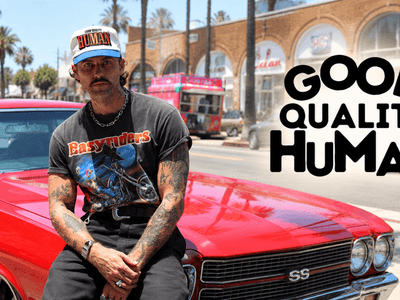 A tattooed man in a vintage t-shirt sits on a red car outdoors