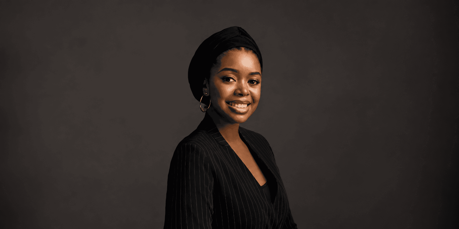 Young woman wearing a black headscarf and pinstripe blazer, smiling against a dark background