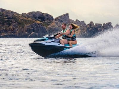 Two riders travel on an Aquamarine jet ski across the Mediterranean with rocky coastline behind them.