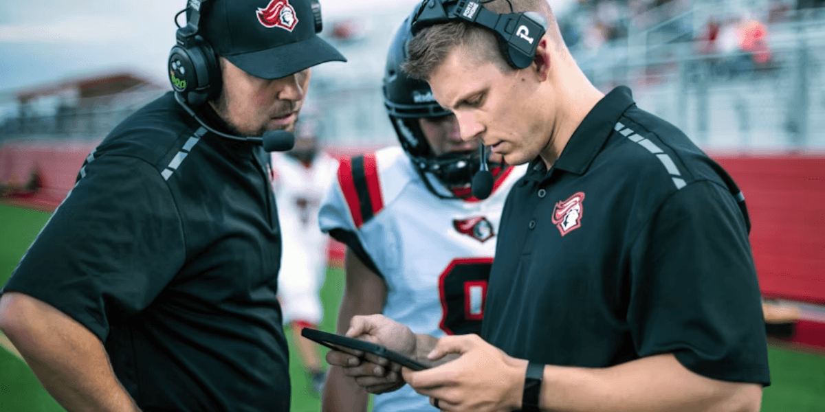 Coaches wearing headsets review a tablet with a football player on the sideline.